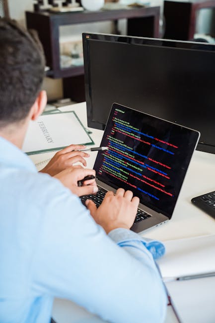 A person seated at a desk working on a laptop displaying lines of colored code. The workspace includes a large monitor, a clipboard with the word 'FEASIBILITY' written on it, and a computer mouse. The