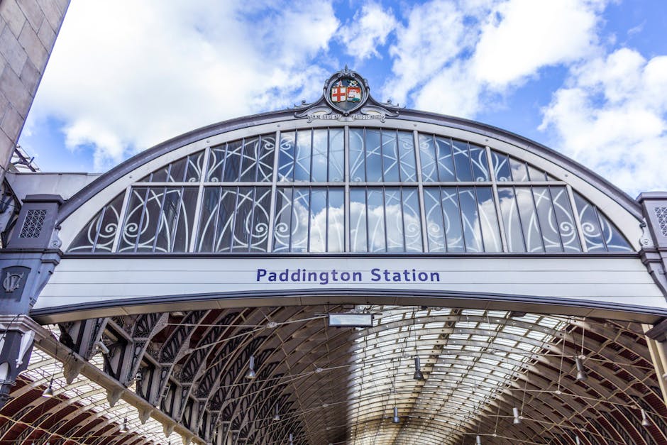 A close-up view of the entrance to Paddington Station showing an ornate, arched metal and glass canopy with intricate scrollwork and decorative elements. The canopy features a crest at its peak with a